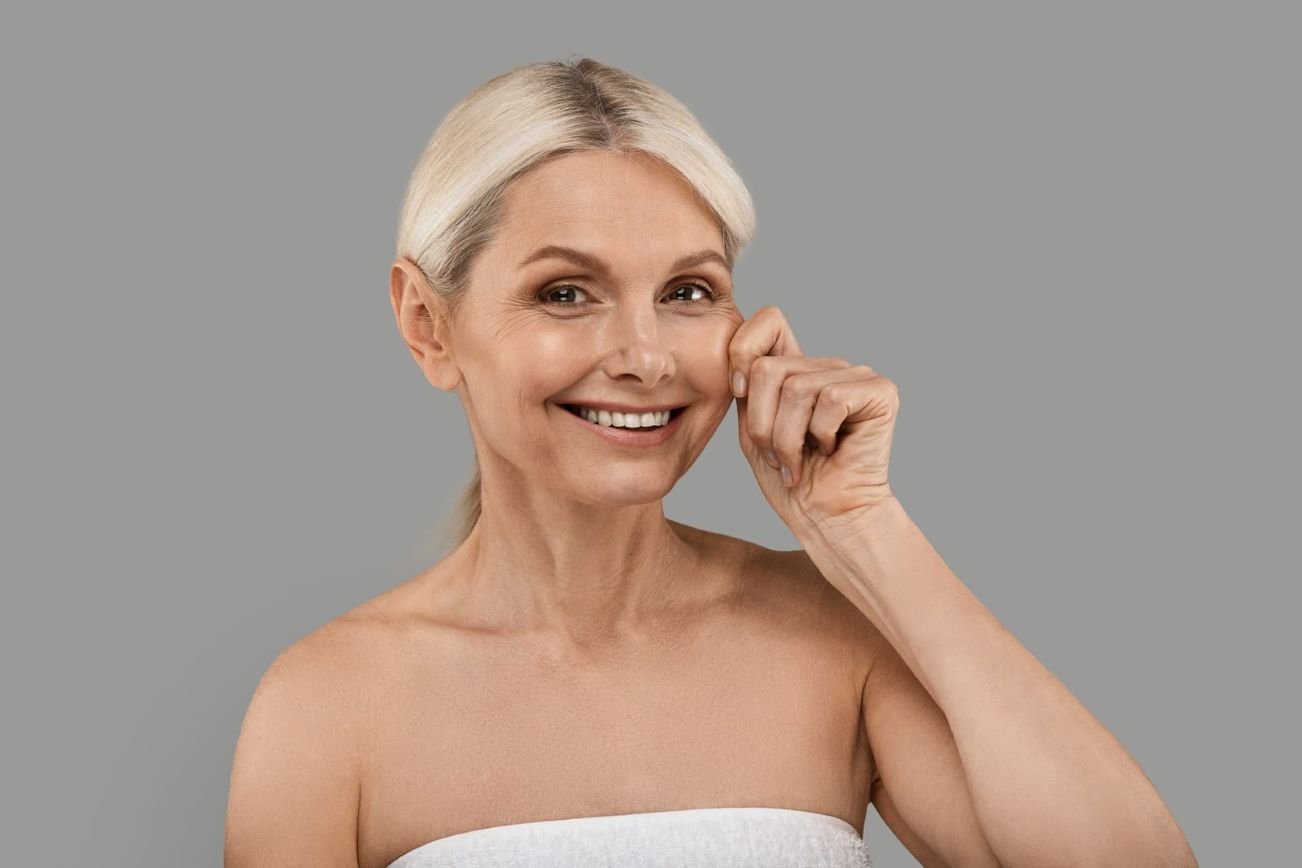 Smiling mature woman with firm, youthful skin pinching her cheek to showcase skin elasticity, representing the benefits of Sublime Skin Firming treatment.