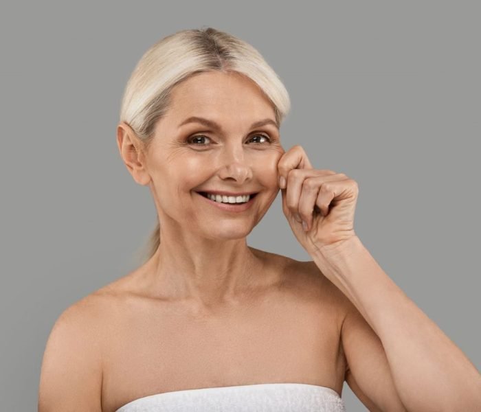 Smiling mature woman with firm, youthful skin pinching her cheek to showcase skin elasticity, representing the benefits of Sublime Skin Firming treatment.