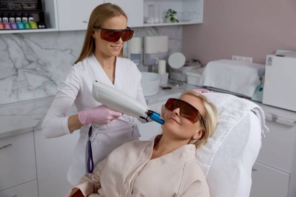 A skincare specialist performing a Pico laser treatment on a woman’s face in a modern aesthetic clinic. Both the specialist and the patient are wearing protective eyewear.